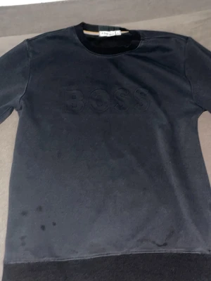 Svart sweatshirt från V-Design - Svart sweatshirt med rund hals och diskret BOSS-tryck framtill. Tröjan har ribbade muddar vid ärmslut och nederkant. Perfekt för en avslappnad och stilren look.