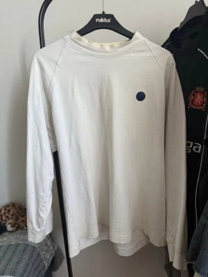 Vit långärmad tröja med blå detalj - Vit långärmad tröja med rund halsringning och en liten blå cirkel som detalj på bröstet. Tröjan har en enkel och clean design, perfekt för en stilren look. Materialet ser ut att vara mjukt och bekvämt, troligen bomull.