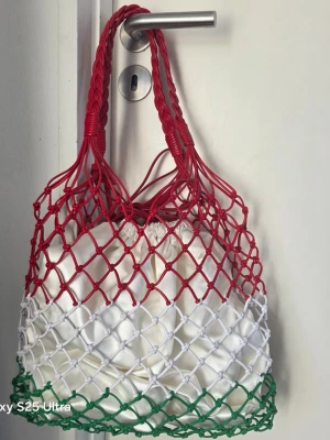 Nätad shoppingväska i rött, vitt och grönt - Säljer en unik shoppingväska med nätmönster i rött, vitt och grönt. Väskan har tvinnade handtag och är tillverkad i slitstark plast. Perfekt för dig som vill ha en färgglad och praktisk accessoar till shoppingen.