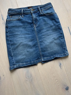 Mörkblå jeanskjol från Lee - Snygg mörkblå jeanskjol från Lee med klassisk femficksdesign, bälteshällor och knappstängning framtill. Kjolen har en rak passform och slits bak för extra rörelsefrihet. Perfekt till sneakers eller boots för en cool look.