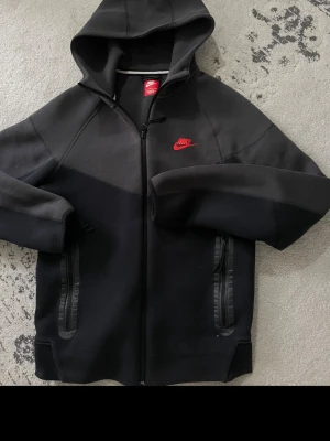 Svart Nike hoodie med dragkedja - Säljer en svart hoodie från Nike med dragkedja framtill och två fickor med svarta detaljer. Hoodien har en röd Nike-logga på bröstet och en stor huva. Materialet är tjockt och mjukt, perfekt för chill dagar. Passar dig som gillar sportig och clean stil. Finns en pyttelitet hål som knappts syns från insidan 