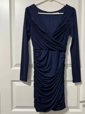 Marinblå omlottklänning med rynk - Snygg marinblå omlottklänning med långa ärmar och rynkade detaljer längs kjolen. Klänningen har en v-ringad omlottdesign och är tillverkad i ett mjukt, stretchigt material som ger en smickrande passform.