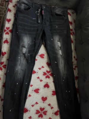 Svarta Dsquared2 jeans med slitningar - Säljer ett par svarta jeans från Dsquared2 med coola slitningar och färgstänk. Jeansen har dragkedja med ringdetalj och klassisk femficksmodell. Snygg streetstil och tydlig Dsquared2-logga vid fickan.