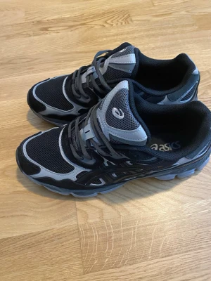 Svarta och grå Asics sneakers - Snygga sneakers från Asics i svart och grått med mesh- och syntetdetaljer. Skorna har klassisk snörning, låg profil och bekväm Ortholite-innersula. Perfekta för dig som gillar sportig stil och vill ha något som funkar till det mesta.