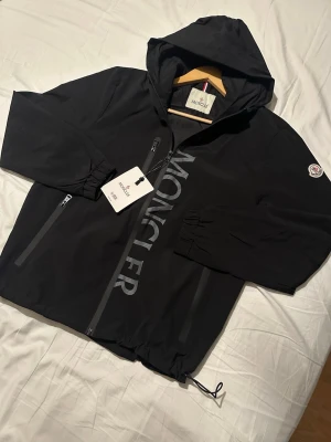 Svart vindjacka från Moncler - Svart vindjacka från Moncler med huva, dragkedja framtill och två sidofickor med dragkedja. Stor Moncler-logga tryckt längs dragkedjan och klassisk patch på ärmen. Jackan har justerbar dragsko nedtill och är tillverkad i lätt, vattenavvisande material.