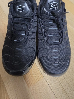 Nike Air Max Plus helsvarta sneakers - Nike Air Max Plus sneakers i helsvart med mesh- och syntetmaterial. Snygga vågformade detaljer på ovandelen, klassisk Nike-logga på tungan och sidan samt robust sula med synliga Air-enheter. Perfekta för dig som gillar streetwear och sportig stil.
