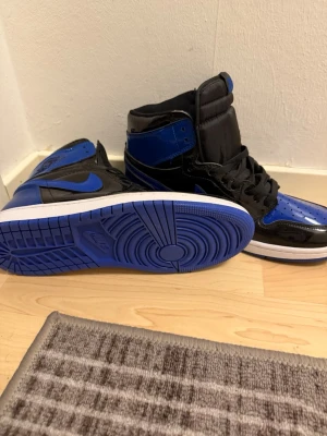 Air Jordan 1 High OG Svart/Blå - Snygga Air Jordan 1 High OG sneakers i svart och blå lackad läder med vit mellansula och blå yttersula. Klassisk siluett med högt skaft, svarta skosnören och ikoniska Swoosh-loggan på sidan. Perfekta för dig som vill sticka ut med en tidlös streetwear-look.