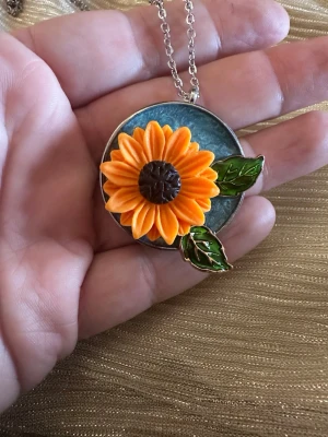 Halsband med solros och gröna blad - Unikt halsband med en stor, detaljerad solros i orange och brunt, samt två gröna blad på en rund blå bakgrund. Hängs i en tunn silverfärgad kedja. Perfekt för dig som gillar färgglada och naturinspirerade accessoarer.