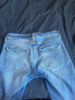 Blå jeans med ficklock - Snygga blå jeans i klassisk modell med raka ben och två bakfickor med lock och knappdetalj. Jeansen har en ljus tvätt och är tillverkade i denim. Perfekta för en avslappnad stil. Liknanade till y2k och true religion. OBS -liten vit prick på knät. kontakta gärna ❤️❤️