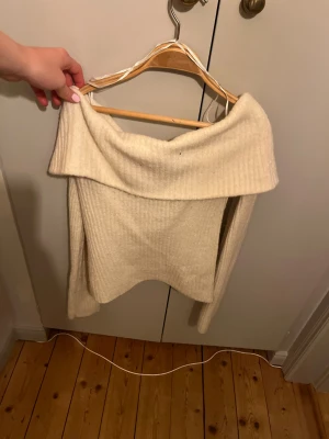 Off shoulder stickad tröja vit/begie - Snygg off shoulder stickad tröja i beige/vit med ribbad struktur. Tröjan har långa ärmar och en bred, nedvikt krage som ger en trendig look. Perfekt för dig som gillar en avslappnad men stilren stil.