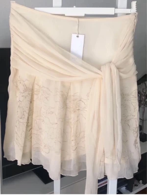 Beige fejk omlottkjol med volangdetalj - Söt kort omlottkjol i ljusbeige chiffong med volang och knytband i midjan. Kjolen har ett skirt lager med diskret blommigt broderi framtill och en flowy känsla. Perfekt för dig som gillar en romantisk och feminin stil. Midjemått 78 cm