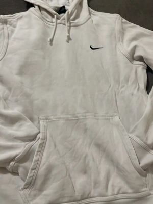 Vit hoodie från Nike med huva - Snygg vit hoodie från Nike med klassisk svart swoosh-logga på bröstet. Hoodien har en stor känguruficka framtill, dragsko i huvan och ribbade muddar vid ärmsluten. Tillverkad i mjukt och bekvämt material, perfekt för en avslappnad stil.