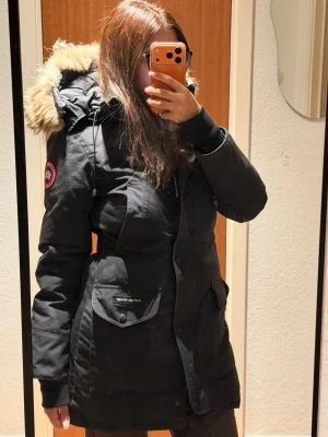 Svart Canada Goose Jacka - Svart parkasjacka från Canada Goose med klassisk patch på ärmen och fuskpäls på huvan. Jackan har dragkedja, stora fickor framtill och justerbar huva. Storlek XS, justerbar i midjan. 