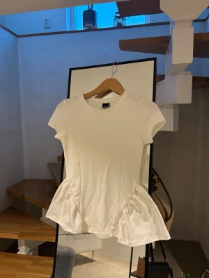 Vit peplumklänning från Gina Tricot - Supersöt vit tröja från Gina Tricot i storlek XS. Modellen har korta ärmar, rund halsringning och en peplumdetalj vid midjan som ger en snygg siluett. Tillverkad i mjukt och stretchigt material för en bekväm passform.
