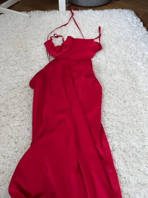 Röd långklänning med smala axelband - Snygg röd långklänning i satinliknande material med smala axelband och v-ringad hals. Klänningen har en elegant och enkel design som ger en lyxig känsla. Perfekt för dig som vill sticka ut med en färgstark look.