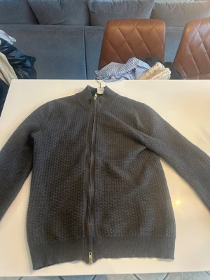 Mörkgrå ziptröja med strukturmönster - Säljer en mörkgrå ziptröja med hög krage och snyggt strukturmönster. Tröjan har hel dragkedja framtill och ribbade muddar vid ärmslut och nederkant. Perfekt för lager på lager och enkel att matcha med olika outfits.John Henric 