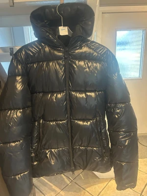 Svart pufferjacka från Save The Duck - Snygg svart pufferjacka från Save The Duck med glansig finish, stor huva och två dragkedjeförsedda fickor framtill. Jackan har ett runt logomärke på ärmen och är tillverkad i syntetmaterial som håller värmen under kalla dagar.