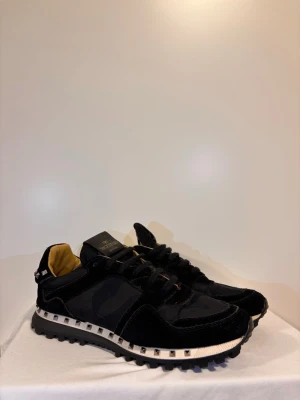Svarta sneakers från Valentino - Snygga svarta sneakers från Valentino med coola nitar runt sulan och en robust yttersula. Skorna har en mix av glansigt och matt material, samt beige innerfoder. Perfekta för dig som vill ha en lyxig och trendig look.