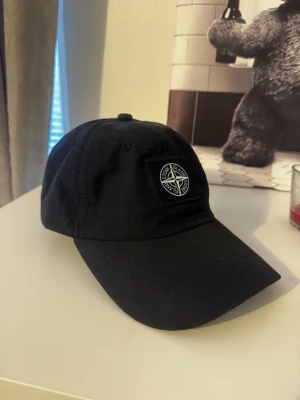 Svart keps från Stone Island - Snygg svart keps från Stone Island med klassisk broderad kompasslogga framtill. Justerbar rem baktill och böjd skärm. Tillverkad i ett lätt och slitstarkt material som passar perfekt till streetwear och casual outfits.