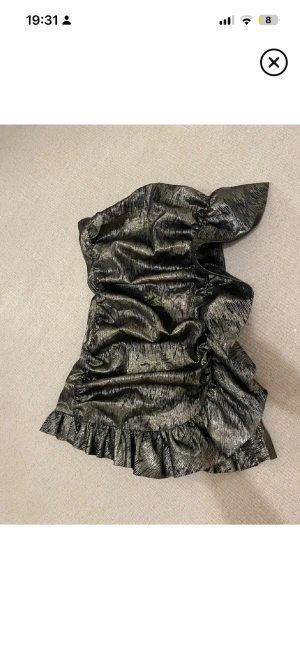 Svart glittrig one shoulder klänning - Snygg svart glittrig kortklänning med one shoulder-design och volangdetalj nertill. Klänningen har en trendig metallic-look och är perfekt för dig som vill sticka ut. Materialet känns lyxigt och skimrande.