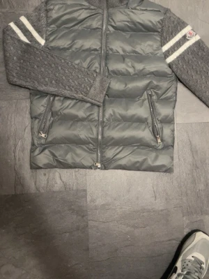 Grå Moncler dunväst med stickade ärmar - Snygg grå dunväst från Moncler med stickade ärmar och vita ränder på överarmarna. Västen har dragkedja framtill, två fickor med dragkedja och Moncler-logga på ärmen. Perfekt för lager-på-lager och streetstyle.