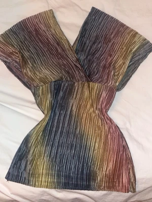 Flerfärgad omlott blus med plissering - Unik omlott blus med plisserat tyg i flera färger som blått, gult, brunt och lila. Blusen har v-ringning och korta ärmar, vilket ger en avslappnad men ändå stilren look. Perfekt för dig som gillar statement-plagg och vill sticka ut.