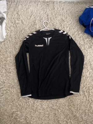 Svart långärmad tröja från Hummel - Svart långärmad sporttröja från Hummel med vita detaljer på axlarna och vid halsen. Tröjan har klassisk Hummel-logga på bröstet och är tillverkad i ett lätt och stretchigt material, perfekt för träning eller match.