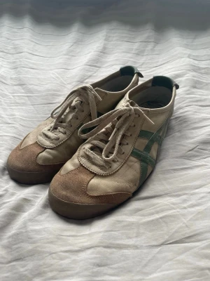 Beige Onitsuka Tiger sneakers med gröna detaljer - Snygga beige sneakers från Onitsuka Tiger med gröna ränder på sidorna och bruna mockadetaljer vid tån. Skorna har klassisk låg profil, snörning och är tillverkade i en mix av textil och mocka. Perfekta för en avslappnad streetstil.