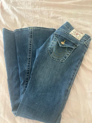  Lowwaist jeans från True Religion - Snygga blå jeans från True Religion (Joey) med klassiska kontrastsömmar och dekorativa fickor bak. Jeansen har låg midja, utsvängda ben och detaljerade knappar. Ej använda mycket och inga defekter, Nypris 1400