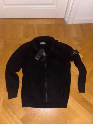 Svart zip-tröja från Stone Island - Säljer en svart stickad zip-tröja från Stone Island med klassisk logopatch på vänster ärm. Tröjan har ribbad struktur, hög krage och dragkedja framtill. Perfekt för dig som gillar streetwear och exklusiva märken.