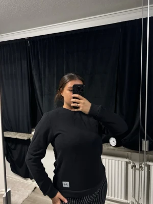 Svart sweatshirt från andy by frank dandy - En enkel och stilren svart sweatshirt från  andy by frank dandy med rund hals och ribbade muddar. Tröjan har en diskret logopatch längst ner på framsidan. Perfekt för dig som gillar klassiska och bekväma plagg.