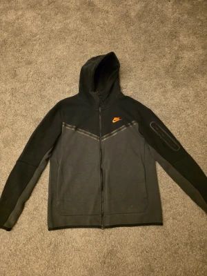 Svart och grå Nike hoodie med dragkedja - Säljer en svart och grå hoodie från Nike med hel dragkedja och huva. Den har ett orange Nike-logo på bröstet och snygga detaljer längs bröstet. Materialet är mjukt och bekvämt, perfekt för chill eller träning. Fickor framtill och cool design på ärmen.