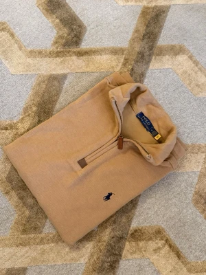 Beige stickad tröja Polo Ralph Lauren - Snygg beige stickad tröja från Polo Ralph Lauren med halv dragkedja och klassisk broderad logga på bröstet. Tröjan har hög krage och ribbade muddar. Perfekt för dig som gillar stilrena och tidlösa plagg.