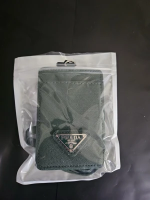 Grön plånbok med axelrem - Stilren grön plånbok i slitstarkt tygmaterial med klassisk trekantig logga i metall framtill. Plånboken har ett fack med transparent fönster på baksidan och en matchande axelrem för smidig användning. Perfekt för kort och småsaker.