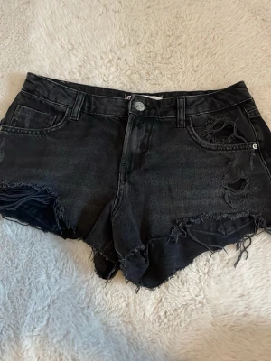 Svarta jeansshorts med slitningar - Snygga svarta jeansshorts från Zara med råa kanter och slitna detaljer framtill. Klassisk femficksmodell med knapp och dragkedja. Perfekta för en avslappnad och trendig look. I storlek 36.