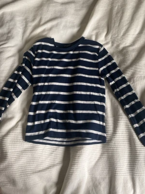 Randig långärmad topp i marinblått - Snygg långärmad topp med marinblå och vita ränder. Klassisk rak passform och rund halsringning. Perfekt att styla med jeans eller kjol för en avslappnad look.