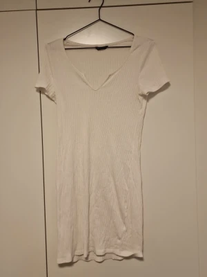 Vit ribbad t-shirtklänning - En enkel och stilren vit t-shirtklänning med ribbad struktur och korta ärmar. Klänningen har en djup v-ringning och är figurnära i modellen. Perfekt för en avslappnad och trendig look.