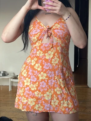 Orange blommig kort klänning med knyt - Snygg kort klänning i orange med blommigt mönster i gult och lila. Klänningen har knyt framtill, v-ringning och är ärmlös med smala axelband. Perfekt för varma dagar och sommarvibbar.