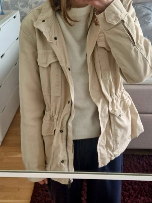 Beige jacka/kappa med fickor - Snygg beige jacka/kappa med dragsko i midjan och flera praktiska fickor framtill. Perkekt nu i vår!🌸 Jackan har lång ärm, knappar och dragkedja. Storlek m/38, men skulle säga att den passar allt från xs-m