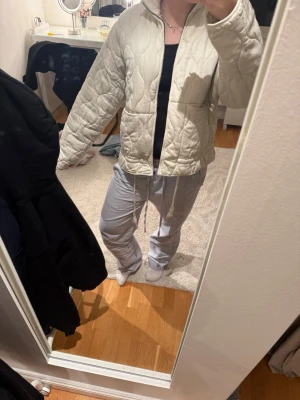Beige quiltad jacka - Snygg beige quiltad jacka med dragkedja framtill och hög krage. Jackan har dragsko i midjan för justerbar passform och två fickor på sidorna. Perfekt för lager-på-lager och en trendig look.