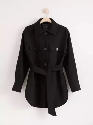 Svart kort damkappa från Lindex - Stilren svart kappa med knappar framtill, två bröstfickor och ett matchande bälte i midjan. Jackan har klassisk krage och långa ärmar med knappdetaljer vid ärmslut. Perfekt för lager-på-lager och enkel att styla till olika outfits.