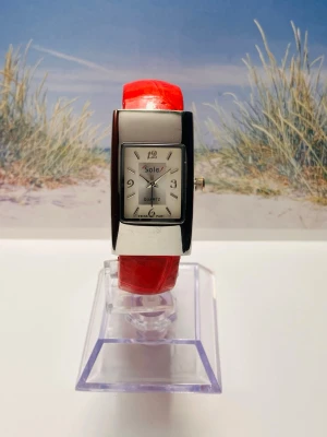 Sole klocka med rött armband - Stilren klocka från Sole med rektangulärt silverfärgat ur och vitt urtavla. Klockan har ett glansigt rött armband som ger en snygg kontrast till det silvriga boetten. Perfekt accessoar för att lyfta din outfit. Nyligen servad med nytt batteri, säljes i befintligt skick enligt bilderna!