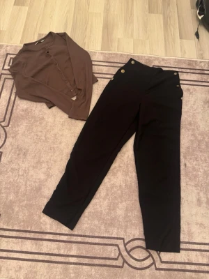 Svarta byxor med knappar i midjan med en brun kofta  - Säljer ett par svarta byxor med snygga guldfärgade knappar i midjan och en brun kofta. Byxorna har en rak passform och hög midja, vilket ger en stilren och trendig look. Perfekta att matcha med en skjorta eller topp för en cool outfit. tröjan funkar som S oxå 