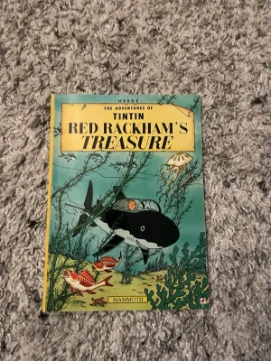 Tintin: Red Rackham's Treasure - Säljer serieboken 'Red Rackham's Treasure' från Tintin-serien av Hergé. Omslaget är färgglatt med undervattensmotiv där Tintin och Haddock utforskar i en ubåt. Boken är i mjukpärm och tryckt på papper. Perfekt för dig som gillar äventyr och klassiska serier.
