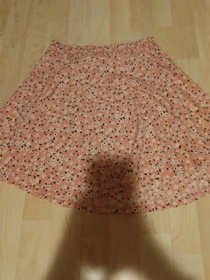 Blommig rosa kjol med resårmidja - Söt kjol i ljusrosa med små vita och svarta blommor. Kjolen har en avslappnad A-linjeform och elastisk midja för skön passform. Perfekt för dig som gillar en feminin och lekfull stil. Materialet känns lätt och luftigt.