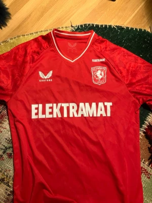 Röd FC Twente matchtröja från Castore - Säljer en röd FC Twente matchtröja från Castore med vita detaljer och klubbmärke på bröstet. Tröjan har sponsortryck från ELEKTRAMAT och THERMO Solutions samt korta ärmar och V-ringad krage. Perfekt för fotbollsfans!