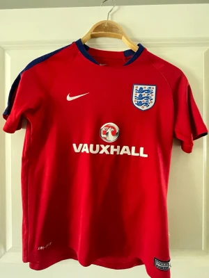 England röd fotbollströja Nike - Röd England fotbollströja från Nike med blå detaljer på axlarna. Tröjan har korta ärmar, broderat landslagsmärke på bröstet och tryckt Vauxhall-logga. Tillverkad i lätt och ventilerande Dri-FIT-material, perfekt för träning eller match.