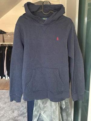 Mörkblå hoodie från Polo Ralph Lauren - Mörkblå hoodie från Polo Ralph Lauren med klassisk känguruficka och huva. Liten röd logga broderad på bröstet. Tillverkad i mjukt bomullsmaterial och har ribbade muddar vid ärmslut och nederkant. Perfekt för en avslappnad och stilren look. Storlek XL (18-20), jag skulle säga att det motsvarar en dam S/M