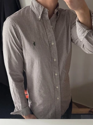 Rutig skjorta från Ralph Lauren - Klassisk rutig skjorta från Ralph Lauren i brunt och vitt. Skjortan har button-down krage, långa ärmar och den ikoniska broderade loggan på bröstet. Tillverkad i mjuk bomull och perfekt för en clean och stilren look.