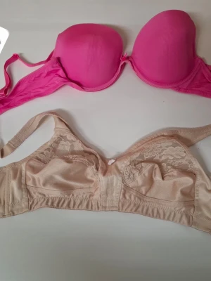 Två bhar i rosa och beige spets - Säljer två bhar, en i starkt rosa med vadderade kupor och bygel, samt en i beige med spetsdetaljer och mjuka kupor utan bygel. Båda har justerbara axelband och hakknäppning bak. Perfekta för dig som vill ha både stöd och komfort.